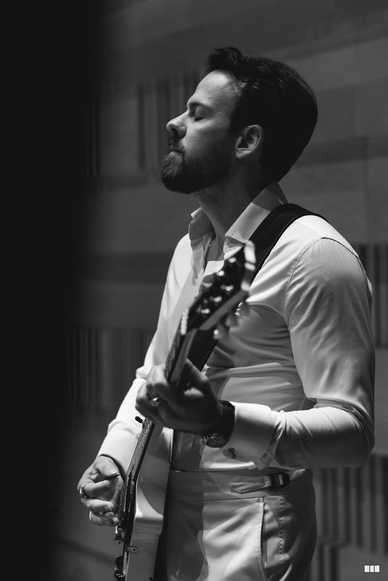 A bearded musician in formal attire plays an electric guitar with eyes closed in focused concentration.