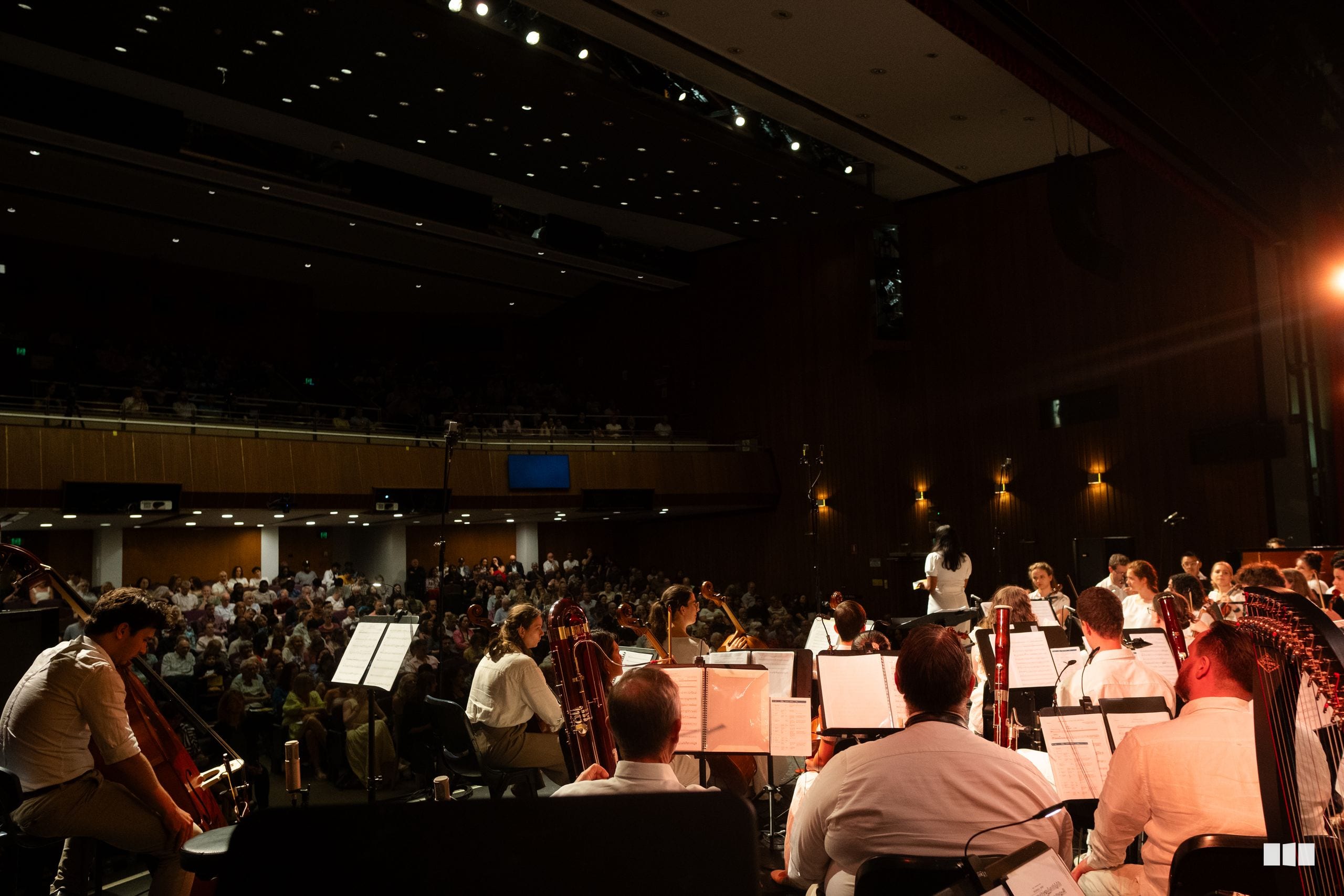 Orchestra musicians perform before a large audience in a concert hall with the conductor leading from the podium.