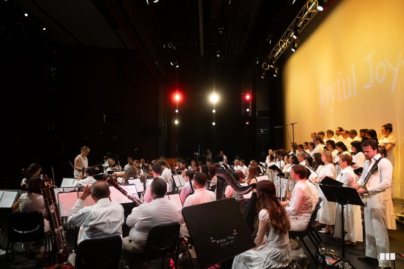 A conductor leads a combined orchestra and choir during a rehearsal or performance on stage with professional lighting.