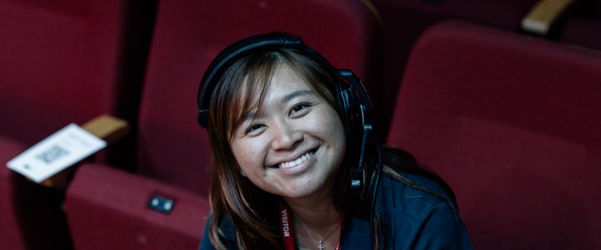 A woman with long brown hair, wearing a headset and a visitor lanyard, seated in a theater with red seats.