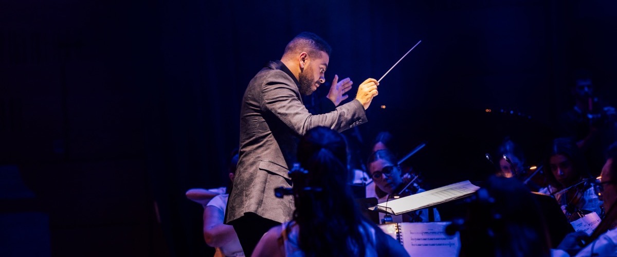 A man in a suit is conducting an orchestra, holding a baton in his right hand.