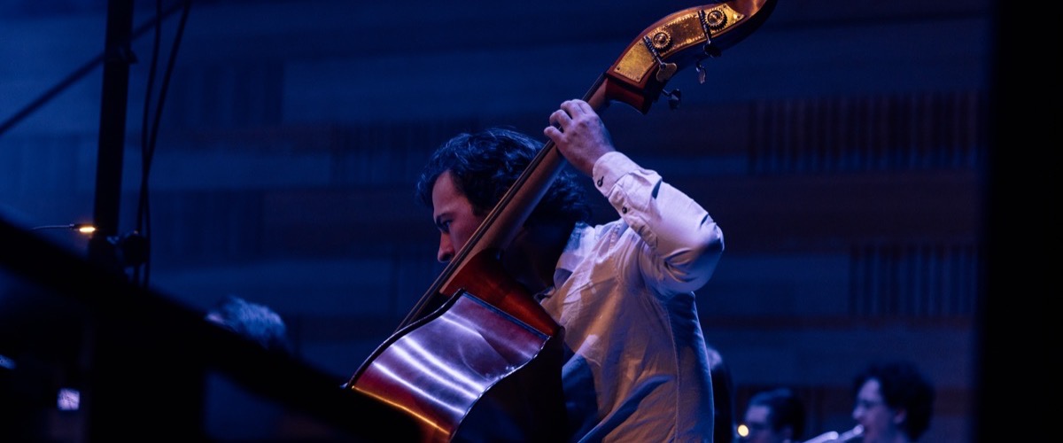 A man with dark hair and a white shirt plays a double bass in a concert setting.