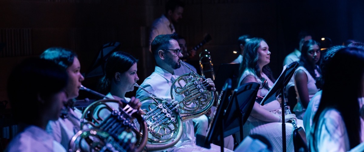 A group of musicians play French horns in a dimly lit, blue-lit concert hall setting.