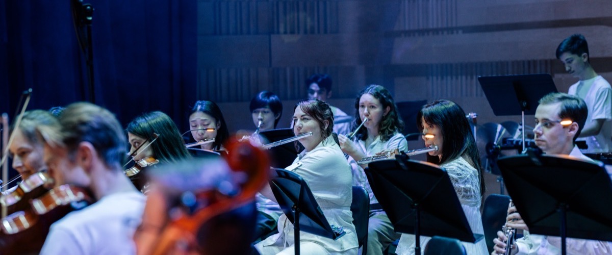 A group of musicians, including flutists and violinists, are seated on stage in a dimly lit performance hall.