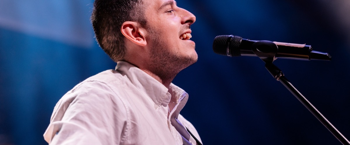 A man is singing into a microphone on a stand, wearing a white collared shirt.