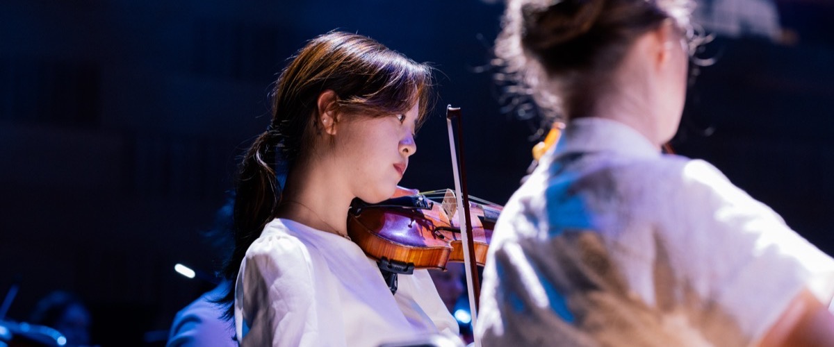 Two young musicians, possibly students, play violins on a dimly lit stage.