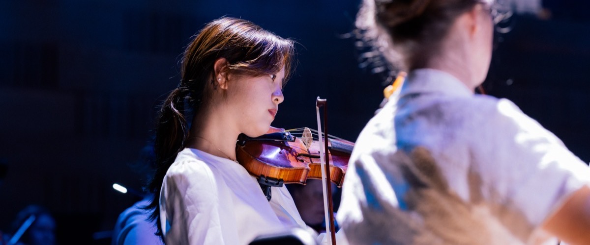 Two people, one playing the violin, are visible from the waist up in a darkened room.