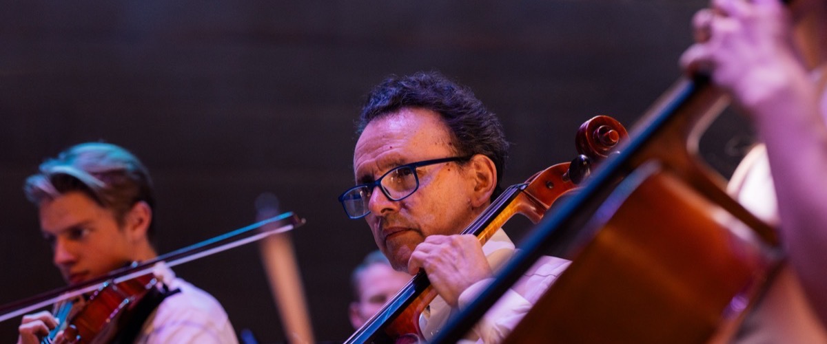 A man playing a cello in an orchestra, with other musicians in the background.