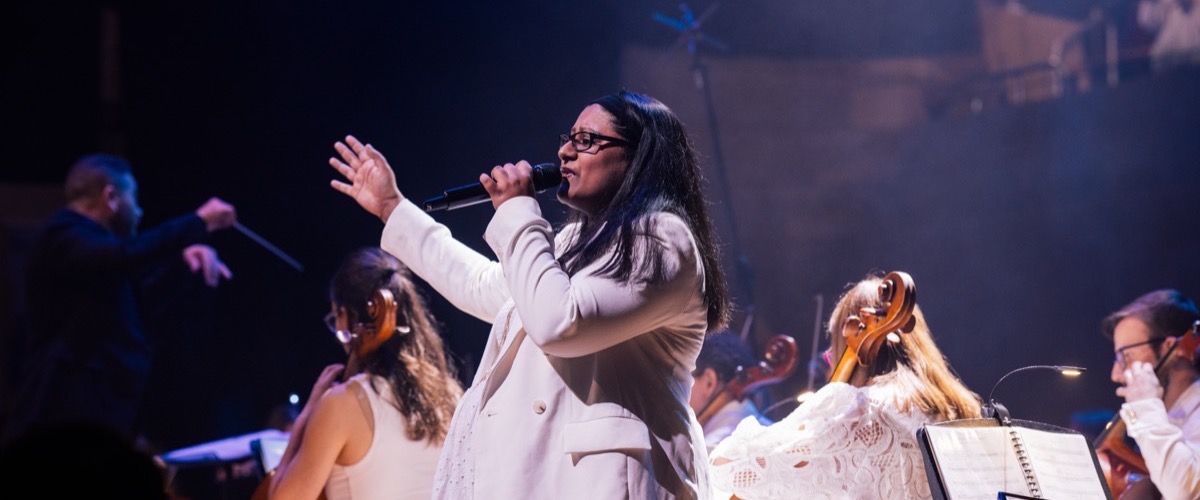 A woman sings into a microphone, accompanied by a conductor and an orchestra.