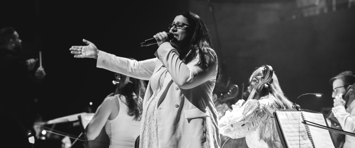 A woman sings into a microphone, accompanied by a group of musicians behind her.