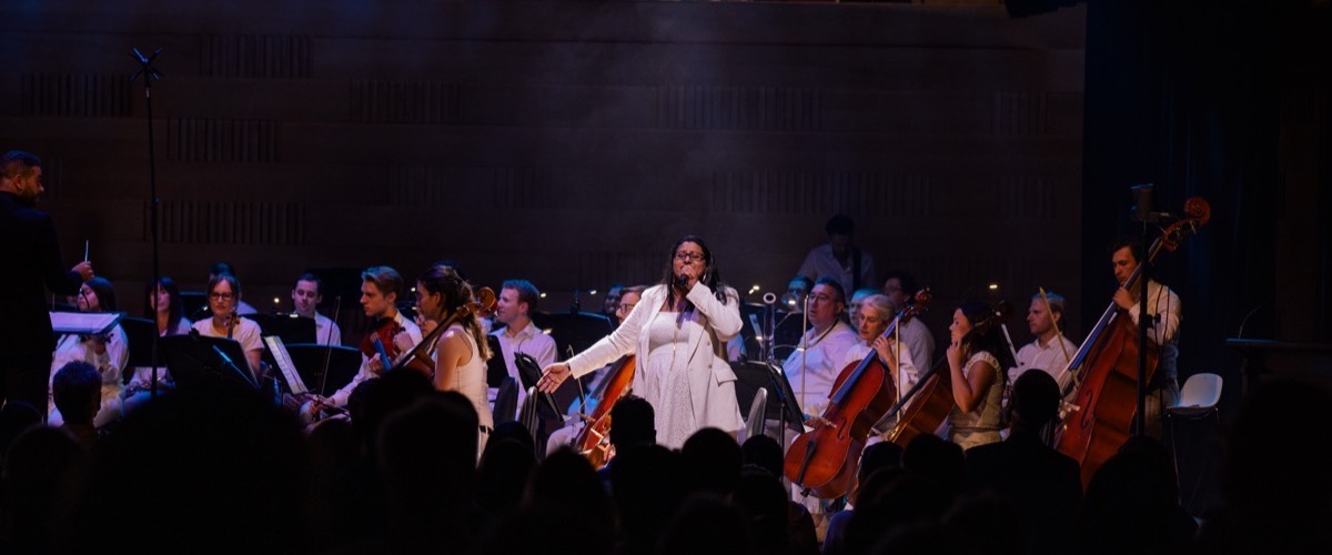 A woman sings while surrounded by a string orchestra, with a conductor leading the ensemble.