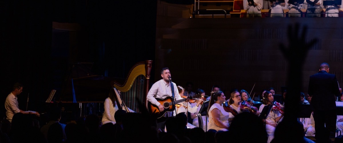 A man plays guitar on stage, surrounded by an orchestra of musicians playing various instruments.