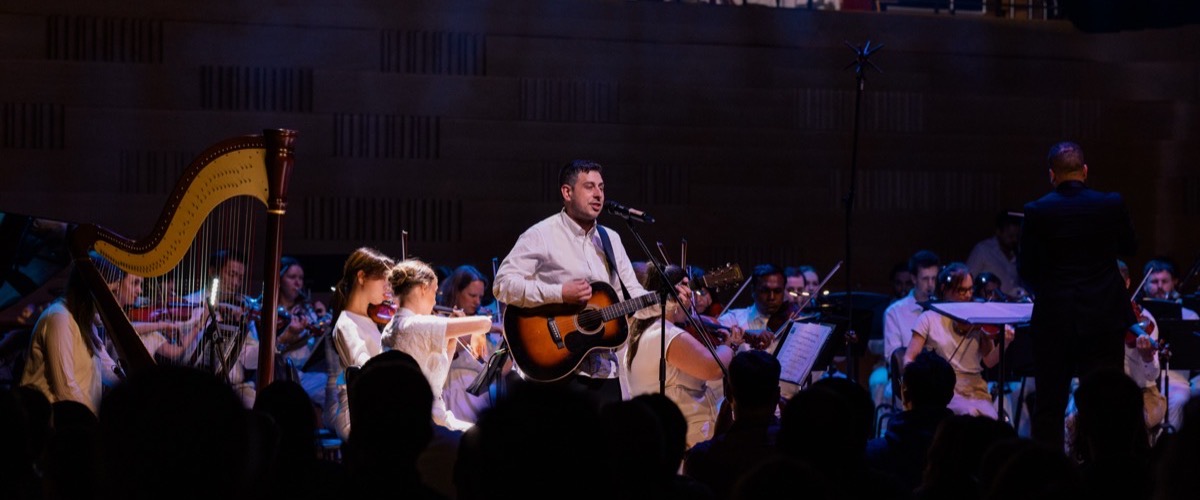 A man playing an acoustic guitar on stage, accompanied by a string orchestra.