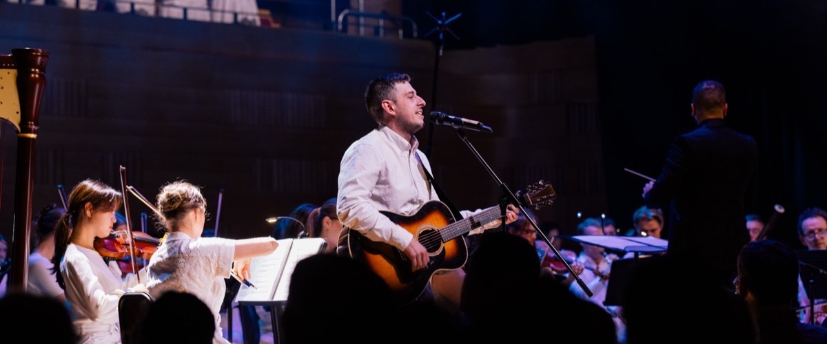 A man singing and playing an acoustic guitar with an orchestra in the background.