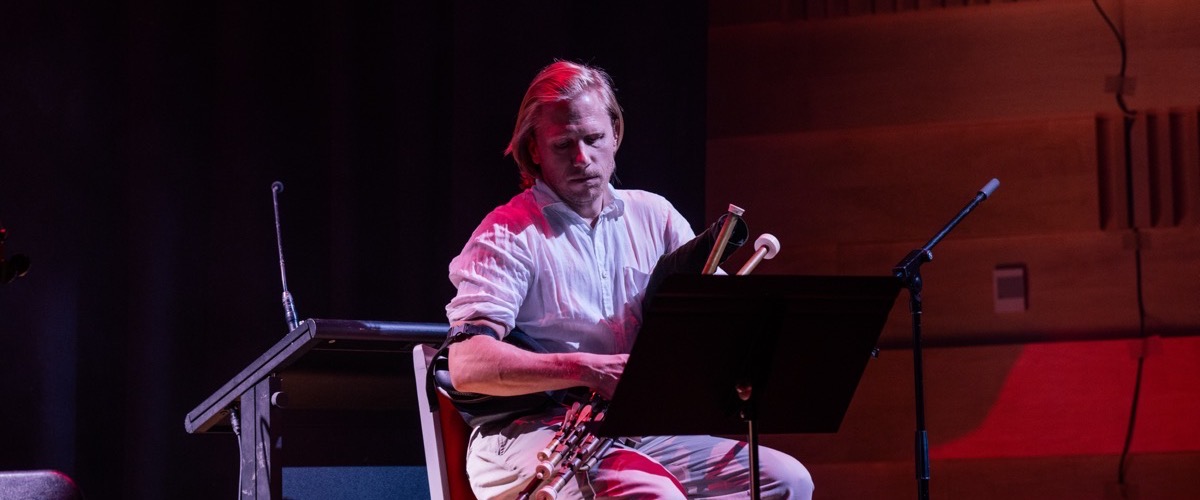 A man seated with a musical instrument, holding a piece of sheet music on a stand in front of him.