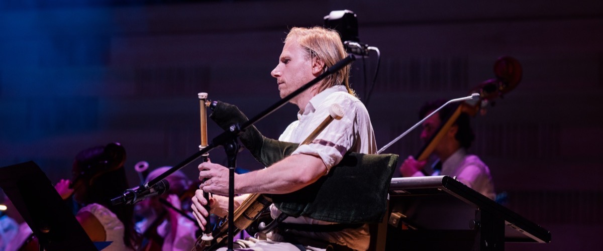 A man with blonde hair plays a bagpipe instrument in front of a microphone and an orchestra.