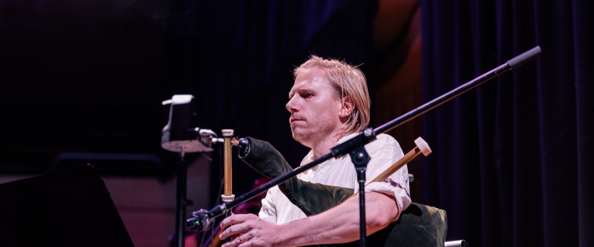 The photograph depicts a man playing a traditional Scottish bagpipe instrument, likely the Great Highland Bagpipe.