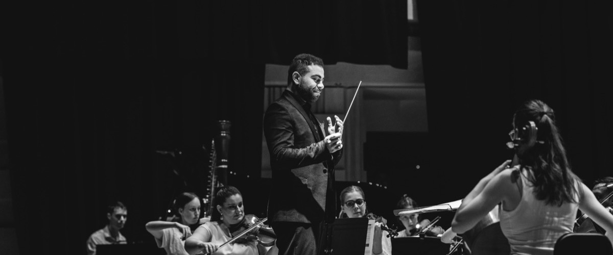 This black-and-white photograph depicts a conductor leading an orchestra, with the conductor holding a baton and the musicians playing various instruments.