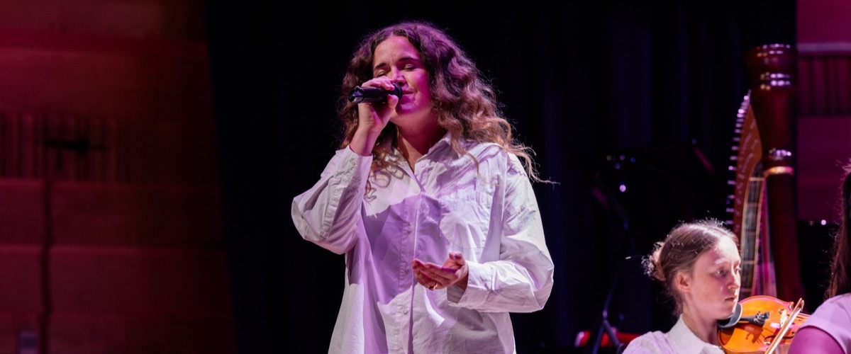 A woman sings into a microphone, accompanied by a musician playing a violin or viola.