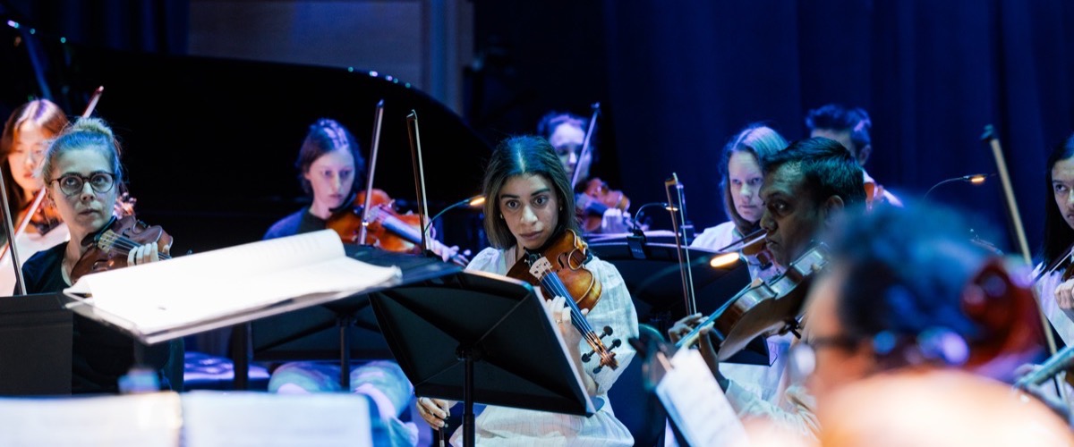 A group of musicians, likely an orchestra, are gathered on stage, playing string instruments.