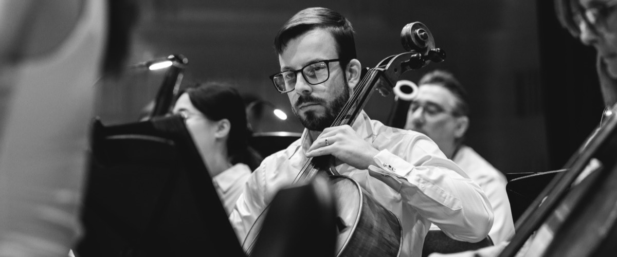 This black-and-white photograph depicts a man playing the cello in a group of musicians.