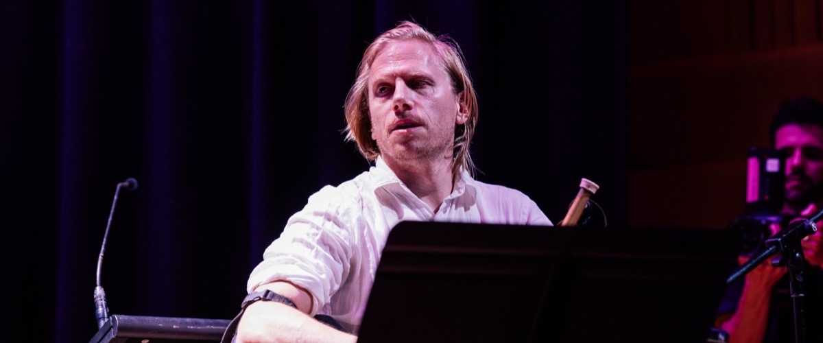 A man with shoulder-length blond hair, wearing a white shirt, seated behind a music stand.