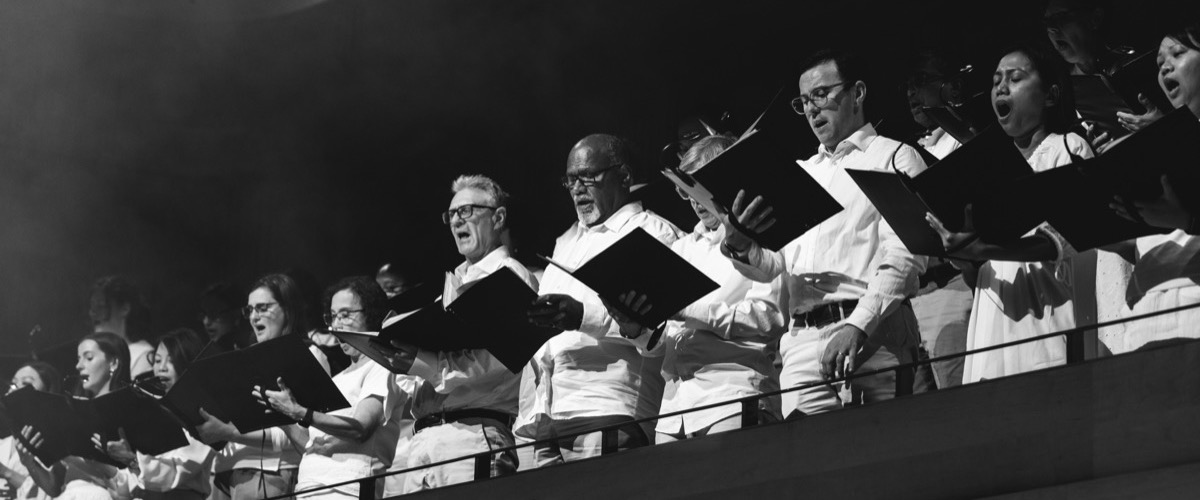 A group of people in white clothing sing together, holding black music sheets.