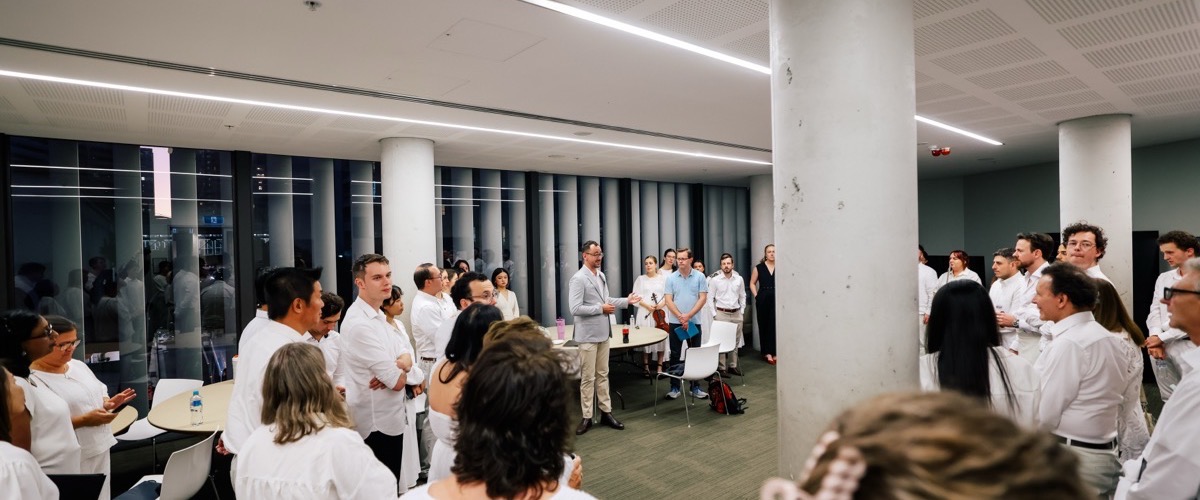 A group of people in a room with white walls and ceiling, standing around tables and pillars.