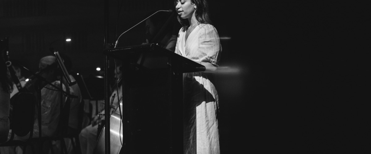 A woman in a white dress stands at a microphone, with a glass podium and dark background.