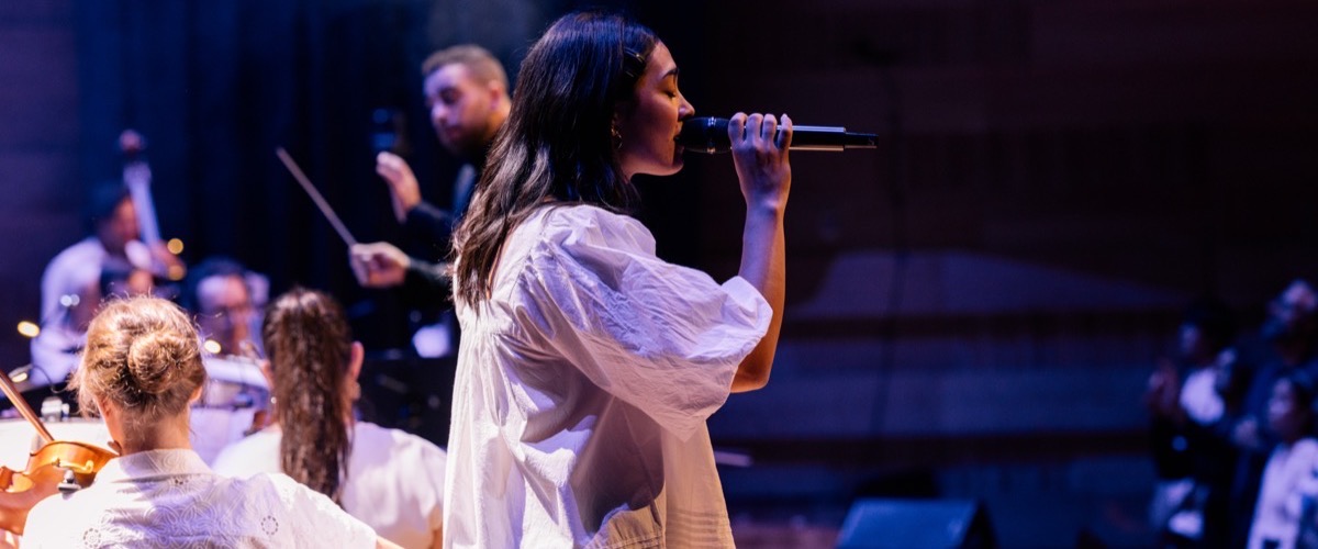 A woman in a white dress singing into a microphone on stage with a string orchestra.