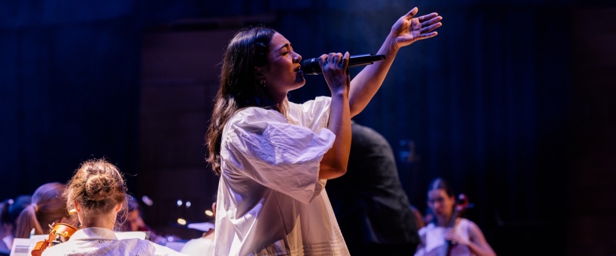 A woman singing into a microphone while a group of musicians play instruments in the background.
