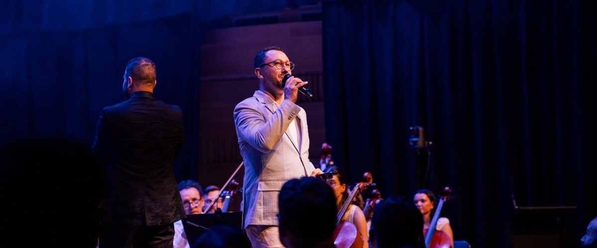 A man singing into a microphone on stage, accompanied by a string section and conductor.