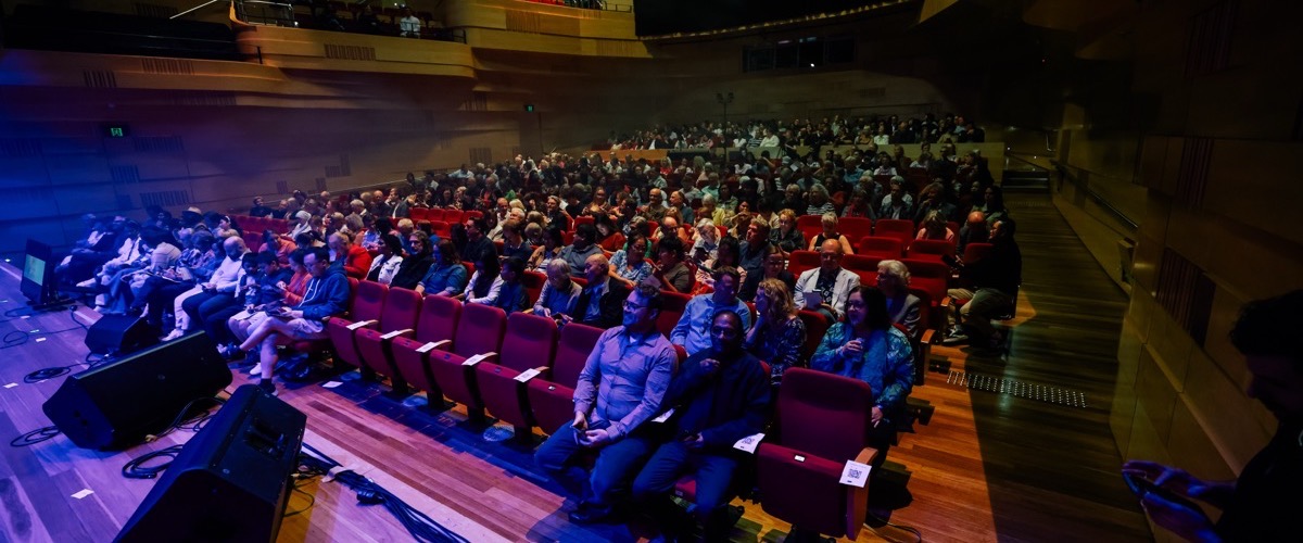 A crowd of people is seated in a large auditorium, awaiting a performance.