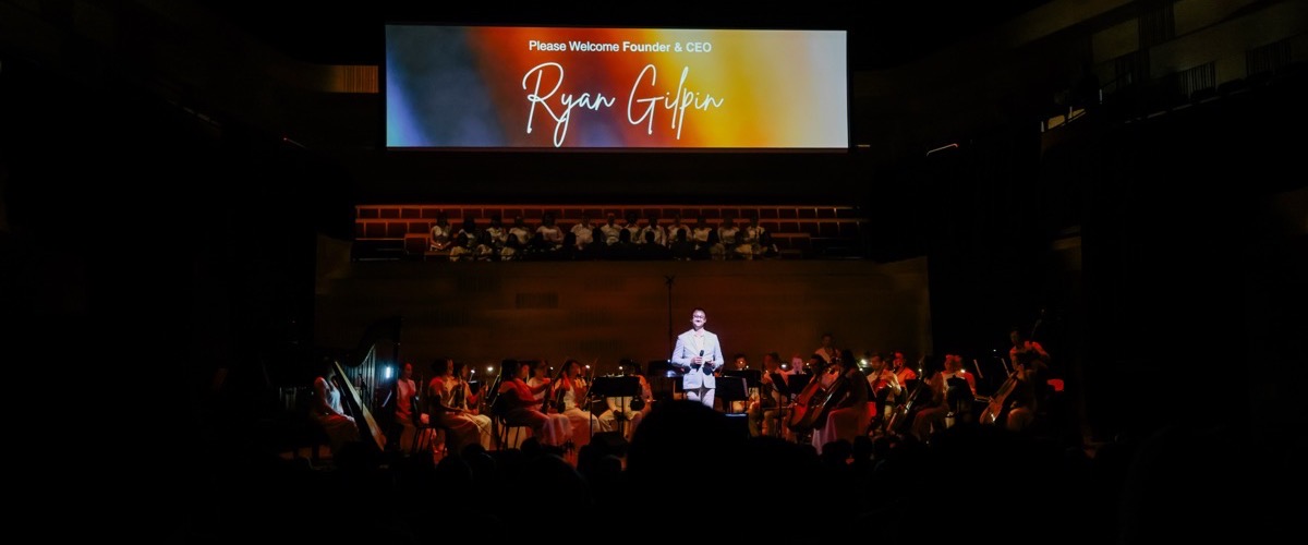 A man standing on a stage in front of an orchestra, with a large screen behind him displaying a welcome message.