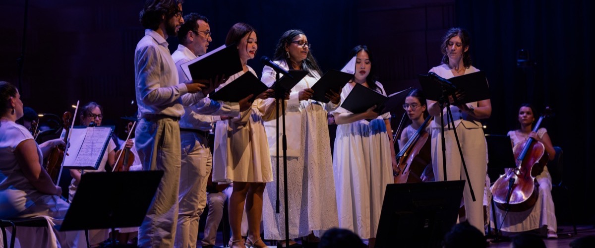 A group of people in white clothing are performing on a stage with sheet music and musical instruments.