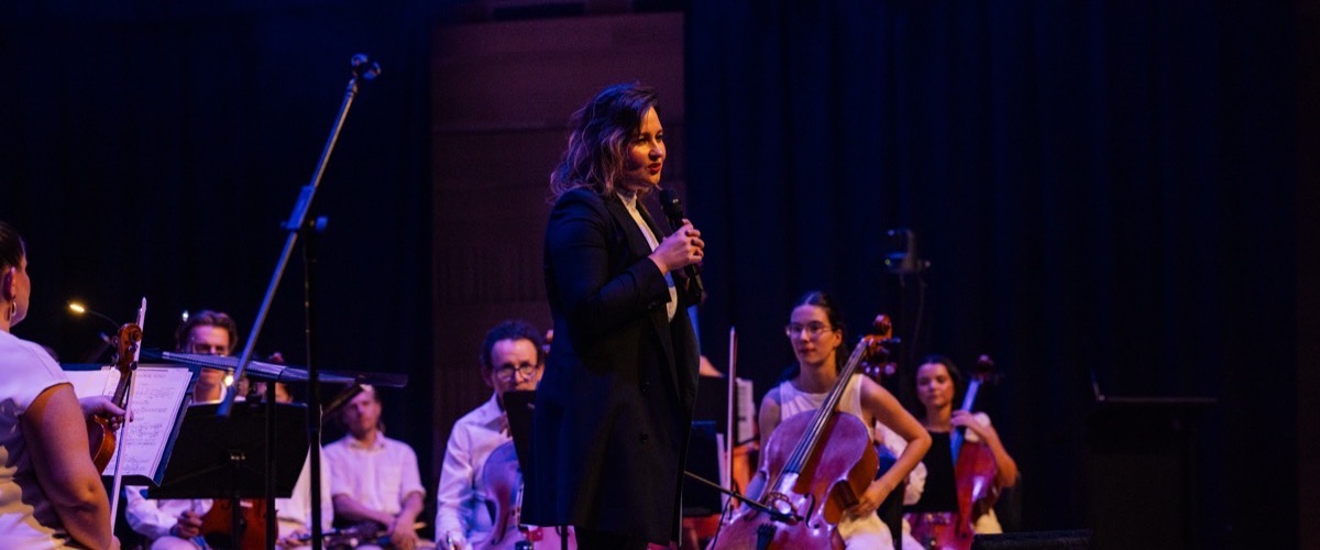 A woman standing in front of an orchestra, speaking into a microphone.