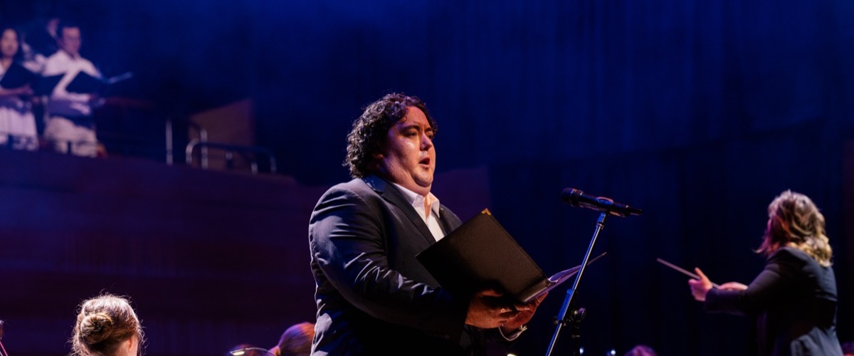 A man singing into a microphone on stage, accompanied by a woman conducting an orchestra.