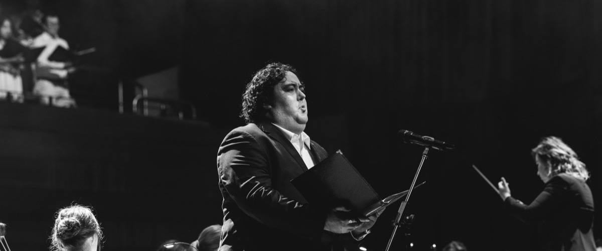 A man in a suit sings into a microphone while holding a music stand and sheet music.