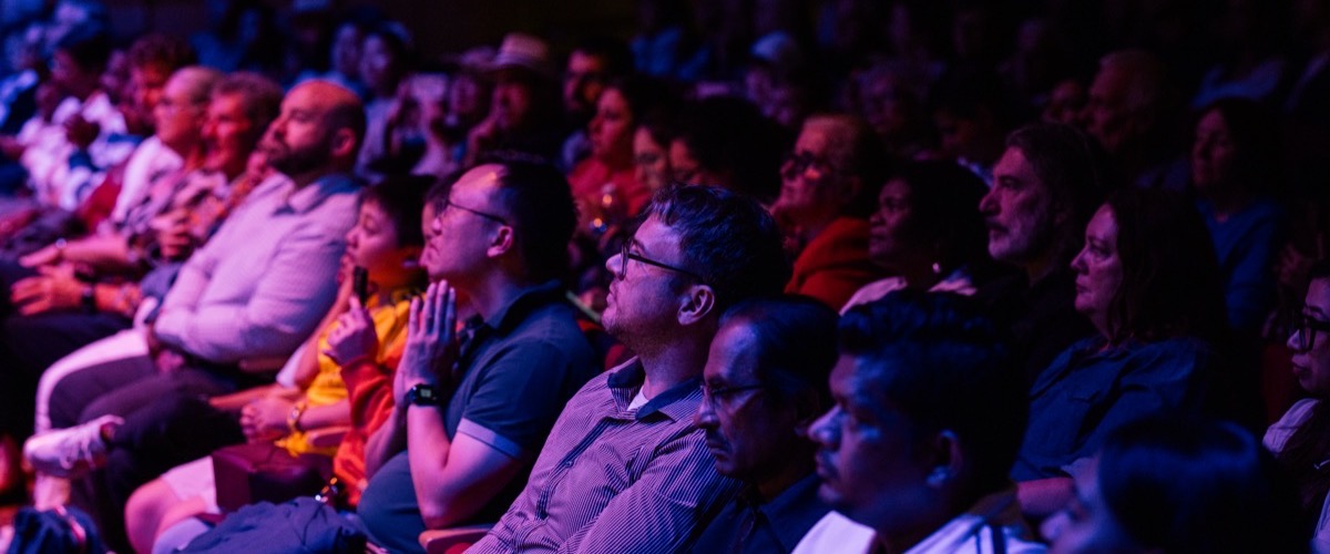 A crowd of people sit in an audience, illuminated by blue and red lighting.