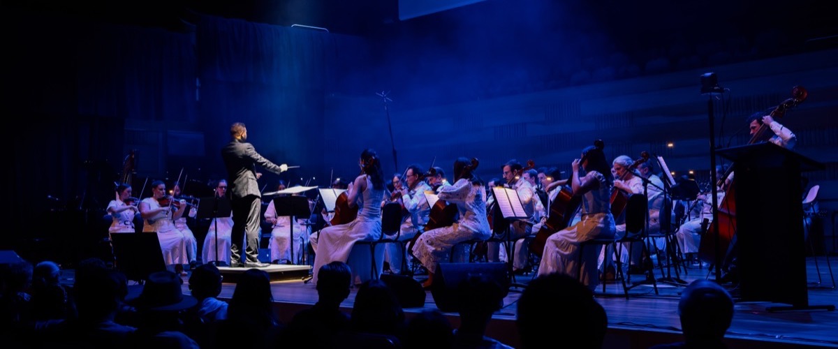 A conductor leads an orchestra of musicians playing string instruments on a stage.
