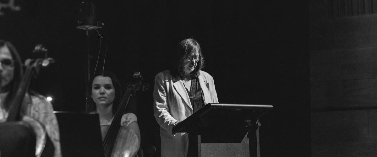 This black and white photograph depicts a woman standing at a lectern, accompanied by two musicians with string instruments.