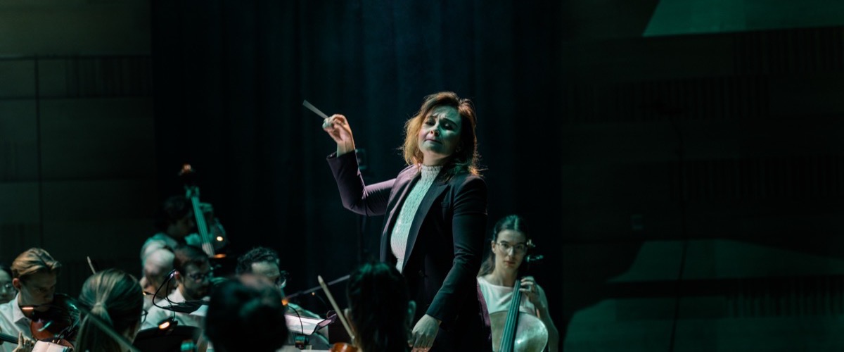 A conductor stands in front of an orchestra, holding a baton aloft in her right hand.