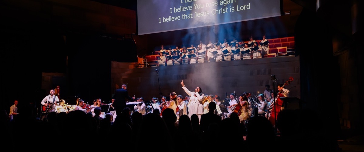 A large group of people performing on stage, with a choir and orchestra, accompanied by a screen displaying lyrics.