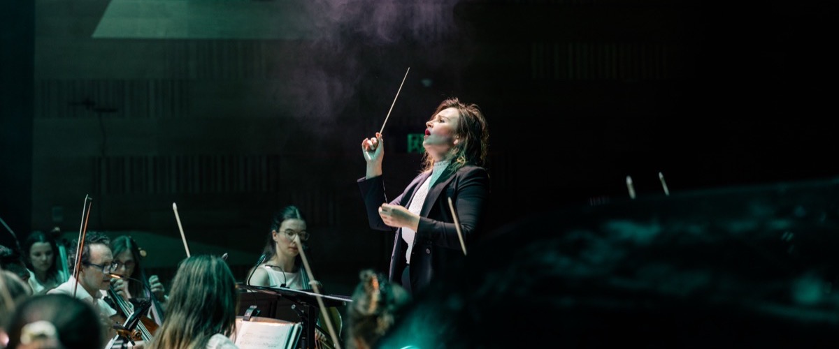 A conductor leads an orchestra, holding a baton aloft, with musicians playing instruments around them.