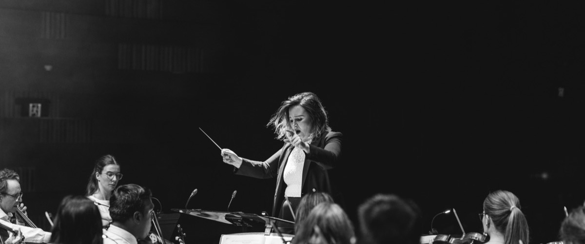 A conductor leads an orchestra, holding a baton and standing before a group of musicians.