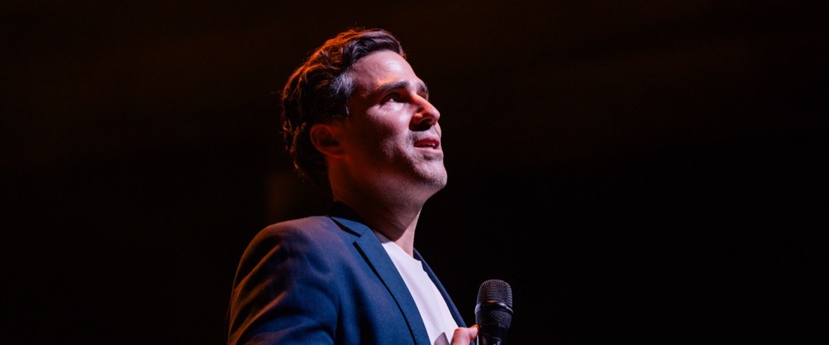 A man in a blue suit jacket holding a microphone on a dark background.