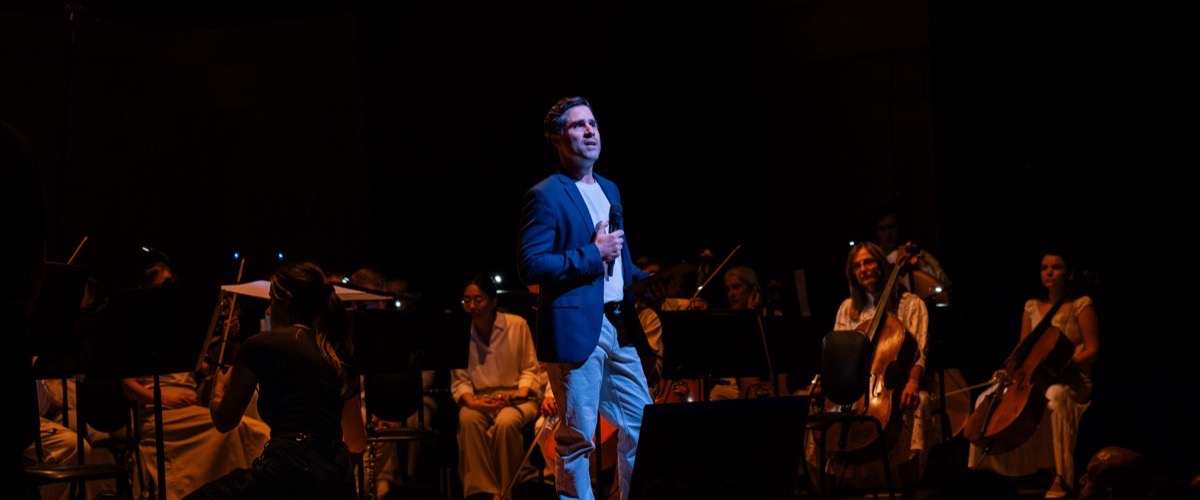 A man in a blue jacket and white shirt sings into a microphone on a stage with an orchestra seated behind him.