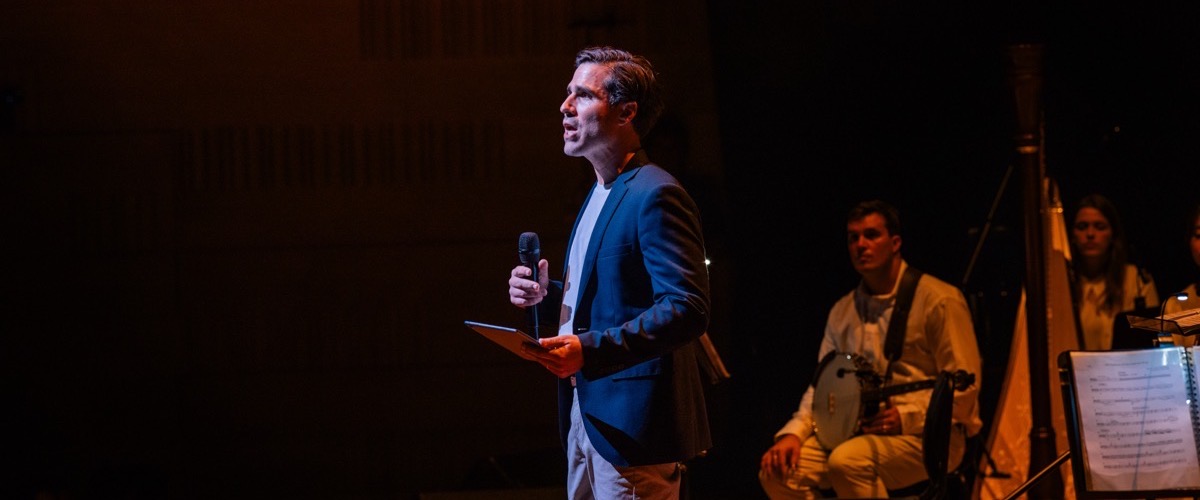 A man in a blue blazer speaking into a microphone on a darkened stage.