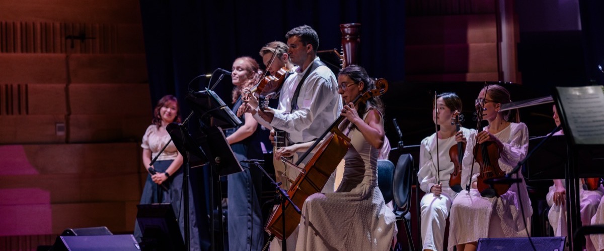 A group of musicians, comprising both men and women, are playing string instruments on a stage.