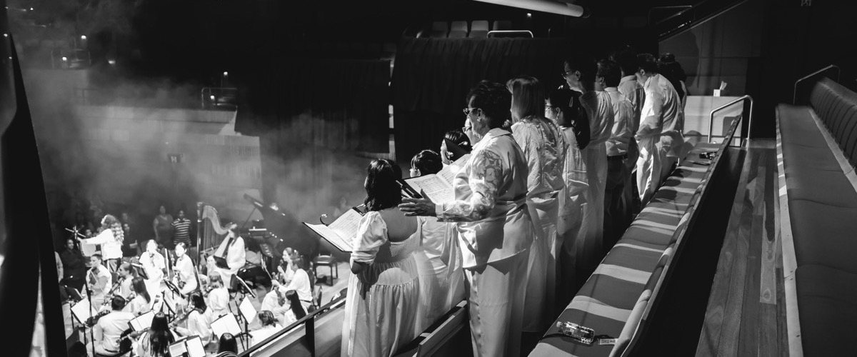 A choir stands in a balcony, overlooking a stage with musicians and other performers.